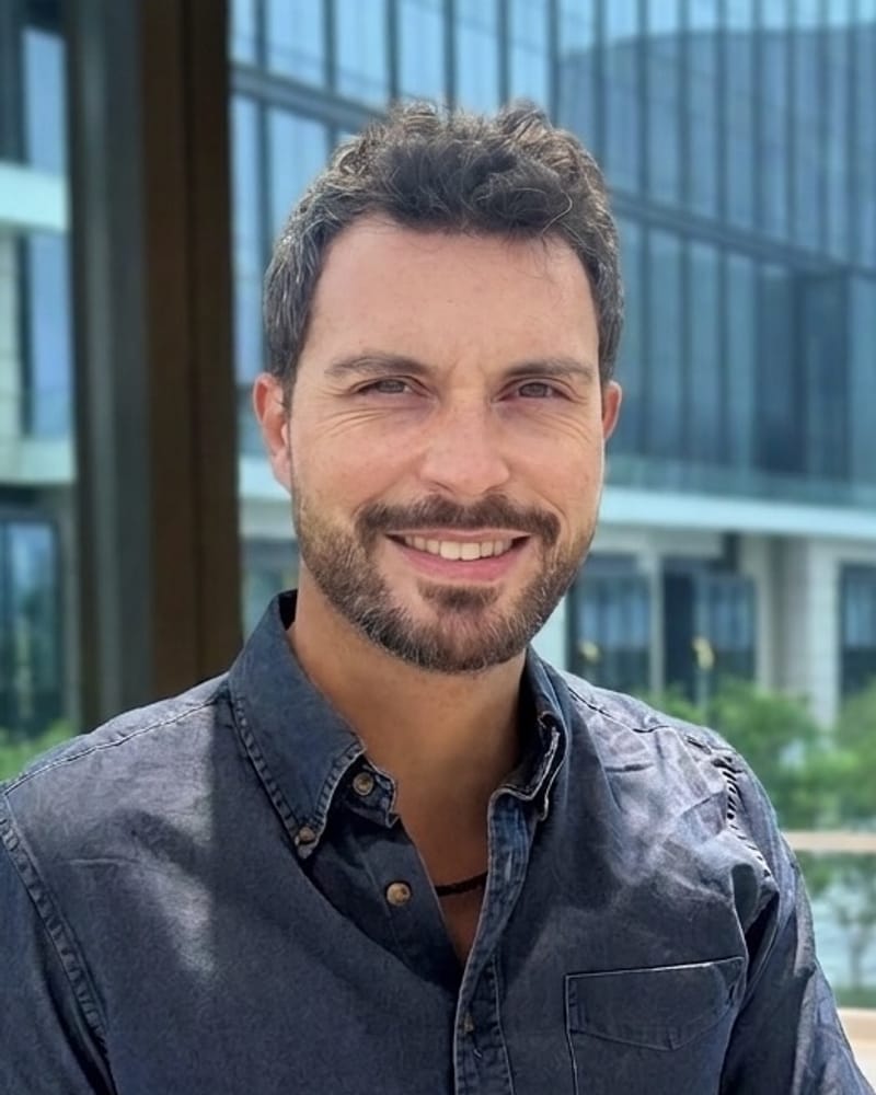 Smiling man with short dark hair and a trimmed beard, in a dark denim shirt, standing outdoors in front of a glass building